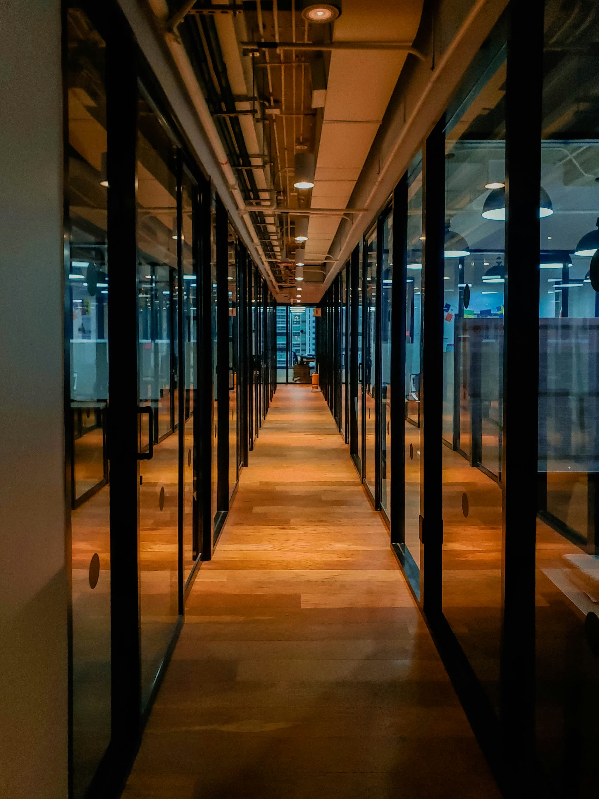A glass-walled office corridor lit warm along a wooden floor.